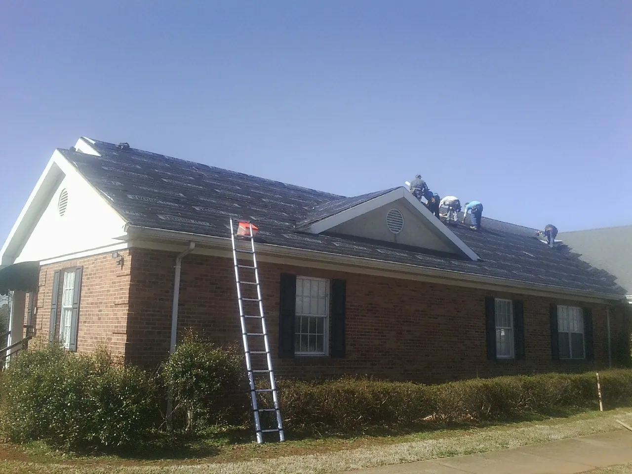 Crew tearing shingles off a brick ranch roof with a ladder set — Emergency Roof Repair work by our crew in Rifton