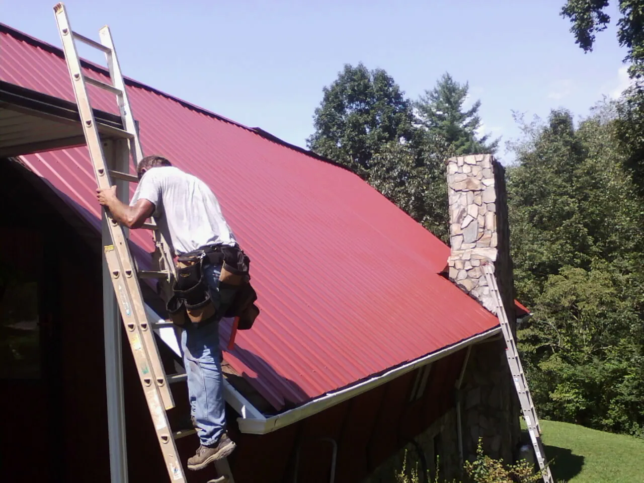 Roofer on a ladder beside a red metal roof and stone chimney in Rifton
